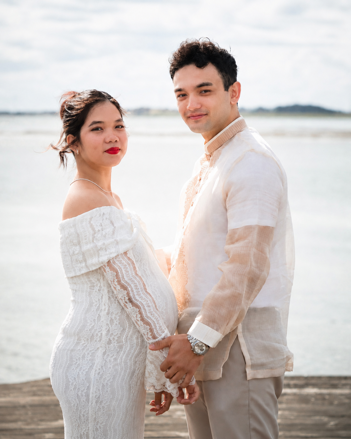 Newlywed couple in elegant light-toned wedding attire standing by the water in Sweden, holding hands and smiling at the camera.