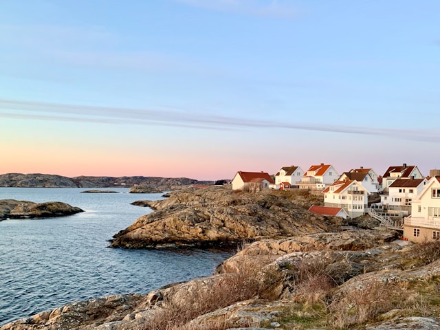 A picturesque coastal village with charming white houses and red roofs nestled on rocky terrain, overlooking a calm sea under a pastel sunset sky. This serene Scandinavian landscape provides a romantic setting, ideal for a unique and intimate wedding experience, perfect for couples seeking natural beauty and tranquility for their special day.
