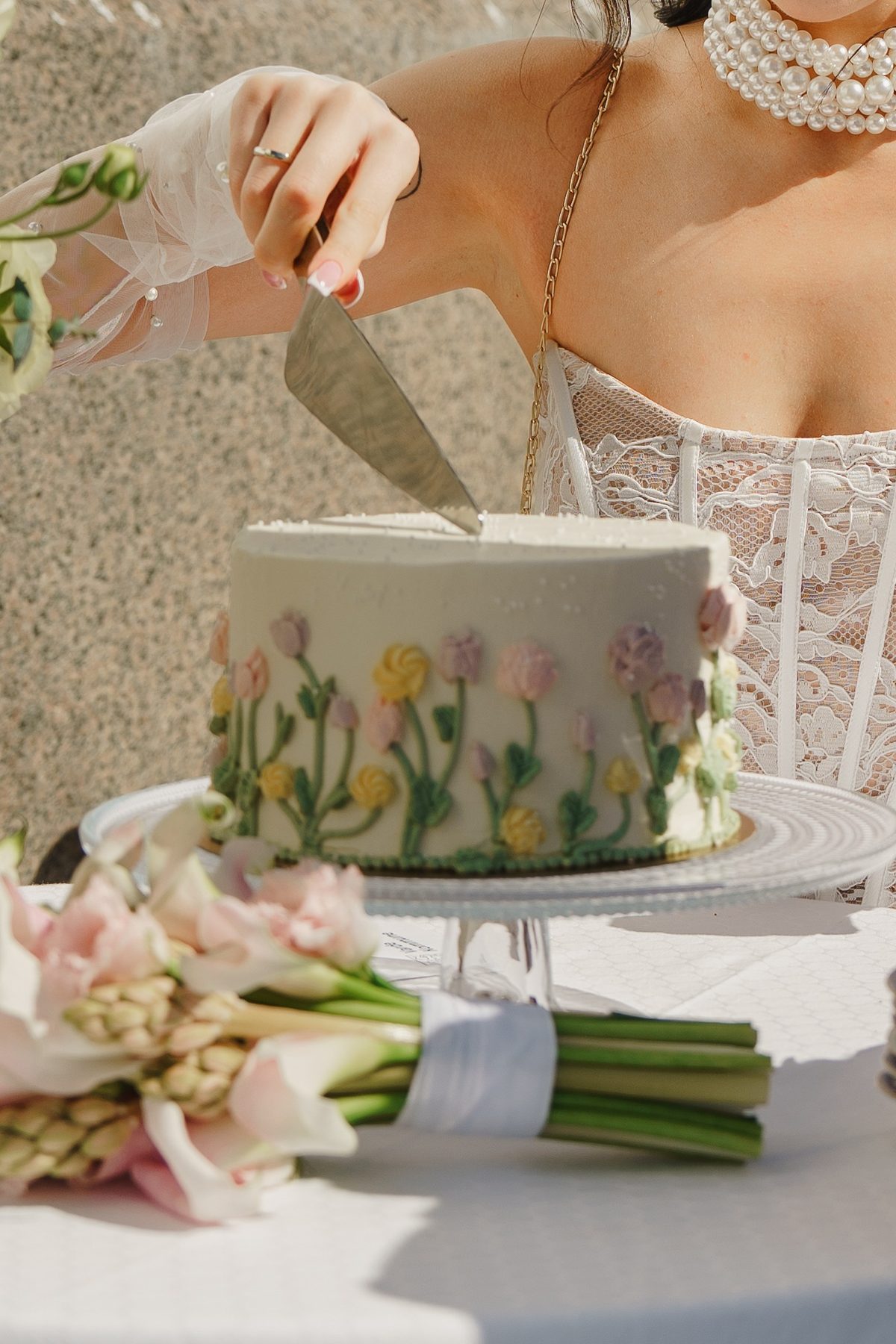 Bride cutting a floral wedding cake beside a pastel bridal bouquet in soft natural sunlight.